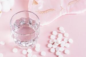 Good sleep concept, sleeping pills, glass of water and sleep mask on pink background photo