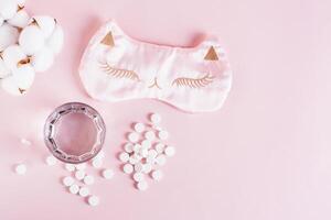 Sleeping pills, a glass of water and a sleep mask on a pink background top view photo