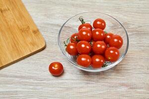 cherry tomatoes in a glass bowl and another one near, on a wooden table surface, near the edge of a cutting board photo