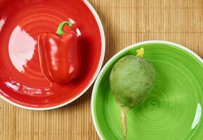 big green radish on a green plate and red bell pepper on a red plate next on a cane place mat photo