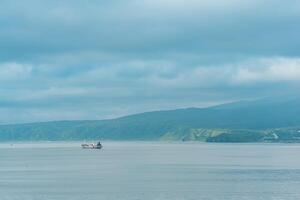 waterscape of the sea bay with a fishing ship and a mount in the clouds in the background, a view of the Mendeleev volcano from the side of the town of Yuzhno-Kurilsk photo