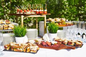 open-air buffet table, glasses and sandwiches on skewers before the start of the holiday against the background of flowering trees in the garden photo