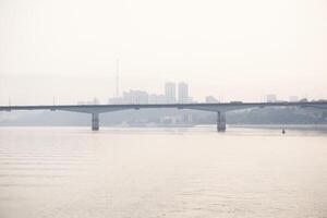 road bridge over a wide river and the silhouette of a distant city behind it in the morning fog photo