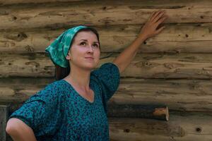 woman in a traditional peasant dress and headscarf dreamily looks at the sky against the background of a log wall photo