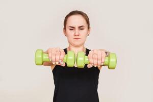 young woman performs exercises with dumbbells, holding them in front of herself with visible effort photo