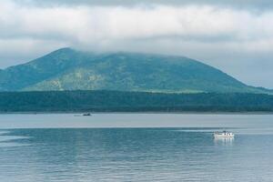 waterscape of the sea bay with a fishing ship and a mount in the clouds in the background, a view of the Mendeleev volcano from the side of the town of Yuzhno-Kurilsk photo
