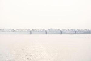 railroad bridge over a wide river and the silhouette of a distant city behind it in the morning fog photo