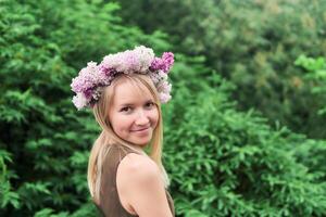 joven mujer en un floral guirnalda de lila flores en un natural antecedentes al aire libre foto