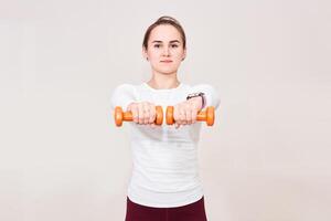 young woman performs exercises with dumbbells, holding them in front of herself, on light background photo