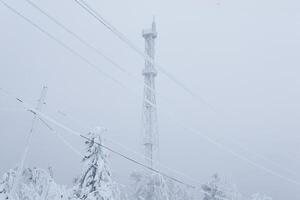 escarchado célula torre y eléctrico alambres terminado un Nevado bosque en parte superior de un montaña en contra un invierno cielo foto