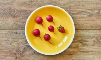 cinco rojo rábanos y uno fresa en un amarillo plato en un de madera mesa foto