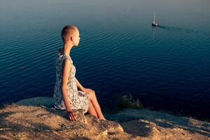 teenager girl in a short haired dress sitting on a cliff over a sea, looking at the ship photo