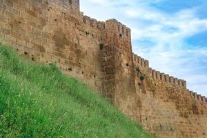 wall of a medieval fortress above a rampart overgrown with grass against the sky, Naryn-Kala citadel in Derbent photo