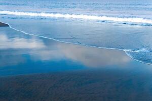 ocean beach with black volcanic sand, the sky is reflected in the rolled back wave of the surf photo