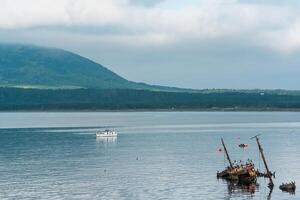 waterscape of the sea bay with a sailing ship and shipwreck and a mount in the clouds in the background, a view of the Mendeleev volcano from the side of the town of Yuzhno-Kurilsk photo