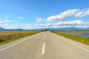 A straight road leading towards mountains under a bright blue sky with clouds, view of lonely road in the ebro delta, tarragona, catalonia, spain photo