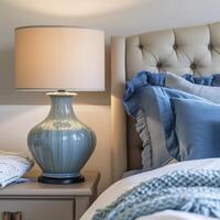 AI generated Close up of blue ceramic lamp on nightstand near bed with beige fabric headboard and blue pillows and blanket photo