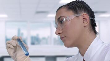 une Jeune femelle scientifique dans une blanc manteau effectue laboratoire tests de une bleu liquide dans une tester tube tandis que séance à une table dans une science laboratoire. fermer planifier.slow mouvement video