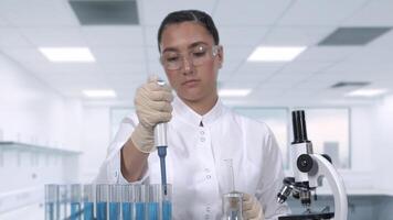 A young female scientist in a white lab coat examines a sample of blue liquid using a micropipette and test tubes sitting at a table in the newest medical laboratory. Slow motion video