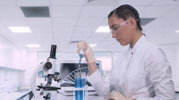 Young female laboratory technician in a white lab coat with dark hair transfers a sample of blue liquid to different test tubes using a micropipette while sitting at a table in a science lab. video