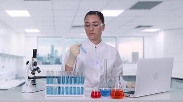 A young female researcher in a white lab coat and goggles examines a liquid in a test tube. A female scientist does medical research and writes the results to a laptop while sitting at a white table. video