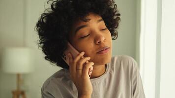 africano americano menina falando em Smartphone às casa interior. jovem mulher com célula telefone conversando com amigos. sorridente adolescente menina fazer respondendo ligar de celular. senhora tendo conversação de Móvel video