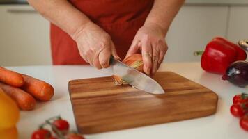 Hands of senior 60s female chopping fresh onion on wooden board while preparing italian pasta with vegetables and soup video