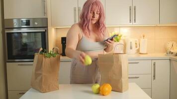 Ordering Food Online. Young pink hair fat lady unpacking grocery bag after shopping, putting vegetables on table in kitchen interior at home video