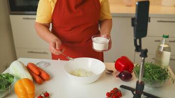 online Koken zelfstudie. Kaukasisch vrouw gebruik makend van slim telefoon voor vloggen bloggen op zoek voor recept terwijl Koken groente veganistisch salade maaltijd. video