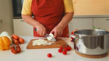 Hands of senior 60s female chopping fresh onion on wooden board while preparing italian pasta with vegetables and soup video