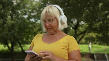 gelukkig oud senior vrouw wandelingen in de park en luistert naar muziek- Aan koptelefoon. een vrouw gepensioneerde is glimlachen en genieten van een warm zomer dag in de tuin. video