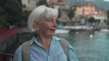 contento 60s anziano donna sorridente per telecamera a lago como paesaggio a soleggiato giorno, femmina godendo sua vacanza vacanza viaggio nel Italia. sorridente femmina turista nel bellissimo paesaggio posto. video
