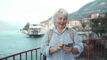 anziano 60s donna viaggiatore utilizzando inteligente Telefono mentre in piedi su banchina contro lago Como, Italia. femmina guardare a Telefono schermo. video