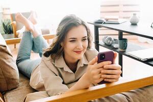 Cheerful Millennial Woman Relaxing on Sofa, Glancing at Phone, Reading Messages, Chatting, and Enjoying Free Time. Cozy Leisure at Home with Happy Young Woman Engaging in Digital Communication. photo