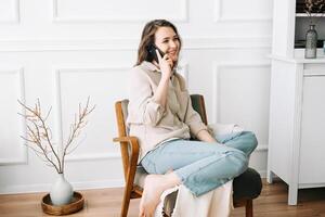 Millennial Woman Smiling, Speaking on Phone, Sitting on Chair, Looking Out Window at Free Space. Smiling millennial female calls by phone, talks, sits on chair looks out window at free space photo