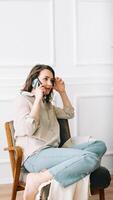 Serious millennial woman talks on the phone, sitting on a chair and looking out the window in her free time. Serious pretty millennial woman calls by phone, talks, sits on chair, looks out window photo