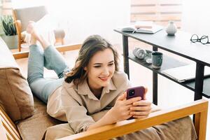 Joyful Millennial Woman Relaxing on Sofa, Engrossed in Phone Messages, Chatting, and Enjoying Leisure Time. Content and Carefree Young Female Lounging on Couch Embracing Digital Communication at Home. photo