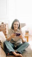 Joyful Millennial Leisure. Smiling young woman indulges in smartphone games, experiencing carefree enjoyment during her free time. Mobile Gaming and Relaxed Tech Fun. photo