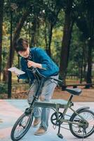 joven hermoso barbado hombre tomando un descanso mientras de viaje el ciudad con su bicicleta utilizando su digital tableta mirando lejos pensativamente foto