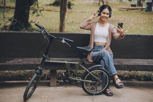 contento joven asiático mujer mientras montando un bicicleta en un ciudad parque. ella sonrió utilizando el bicicleta de transporte. ambientalmente simpático concepto. foto
