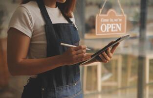 Startup successful small business owner sme beauty girl stand with tablet smartphone in coffee shop restaurant. Portrait of asian tan woman barista cafe owner. SME entrepreneur seller business concept photo