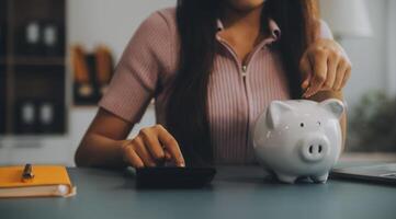 joven asiático mujer ahorro para Jubilación ahorro dinero mediante un cerdito banco y tomando notas en computadora portátil, ahorros concepto. foto