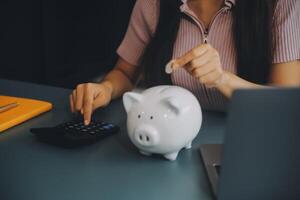 joven asiático mujer ahorro para Jubilación ahorro dinero mediante un cerdito banco y tomando notas en computadora portátil, ahorros concepto. foto