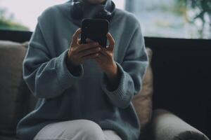 Happy pretty millennial Indian girl relaxing at home, resting in armchair, typing on smartphone, using online app, software, shopping on Internet, making video call. Mobile phone communication photo