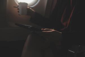 Blonde female tourist checking incoming notification on smartphone sitting on seat of airplane with netbook.Young businesswoman share media from telephone on laptop computer during plane flight photo