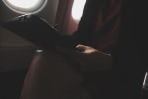 Blonde female tourist checking incoming notification on smartphone sitting on seat of airplane with netbook.Young businesswoman share media from telephone on laptop computer during plane flight photo