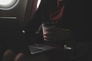 Blonde female tourist checking incoming notification on smartphone sitting on seat of airplane with netbook.Young businesswoman share media from telephone on laptop computer during plane flight photo