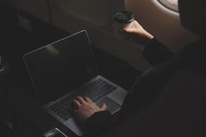 Blonde female tourist checking incoming notification on smartphone sitting on seat of airplane with netbook.Young businesswoman share media from telephone on laptop computer during plane flight photo