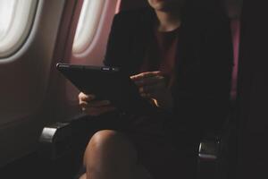 Blonde female tourist checking incoming notification on smartphone sitting on seat of airplane with netbook.Young businesswoman share media from telephone on laptop computer during plane flight photo
