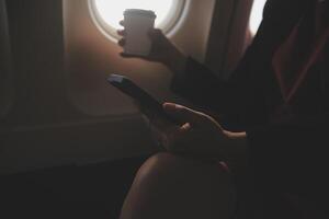 Blonde female tourist checking incoming notification on smartphone sitting on seat of airplane with netbook.Young businesswoman share media from telephone on laptop computer during plane flight photo